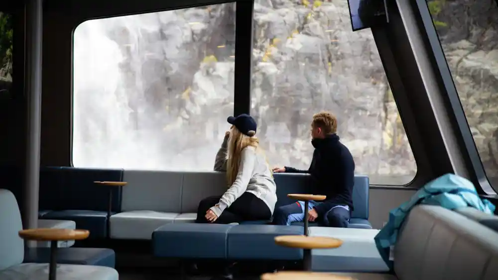 Two people sitting inside boat cabin viewing cliff waterfall